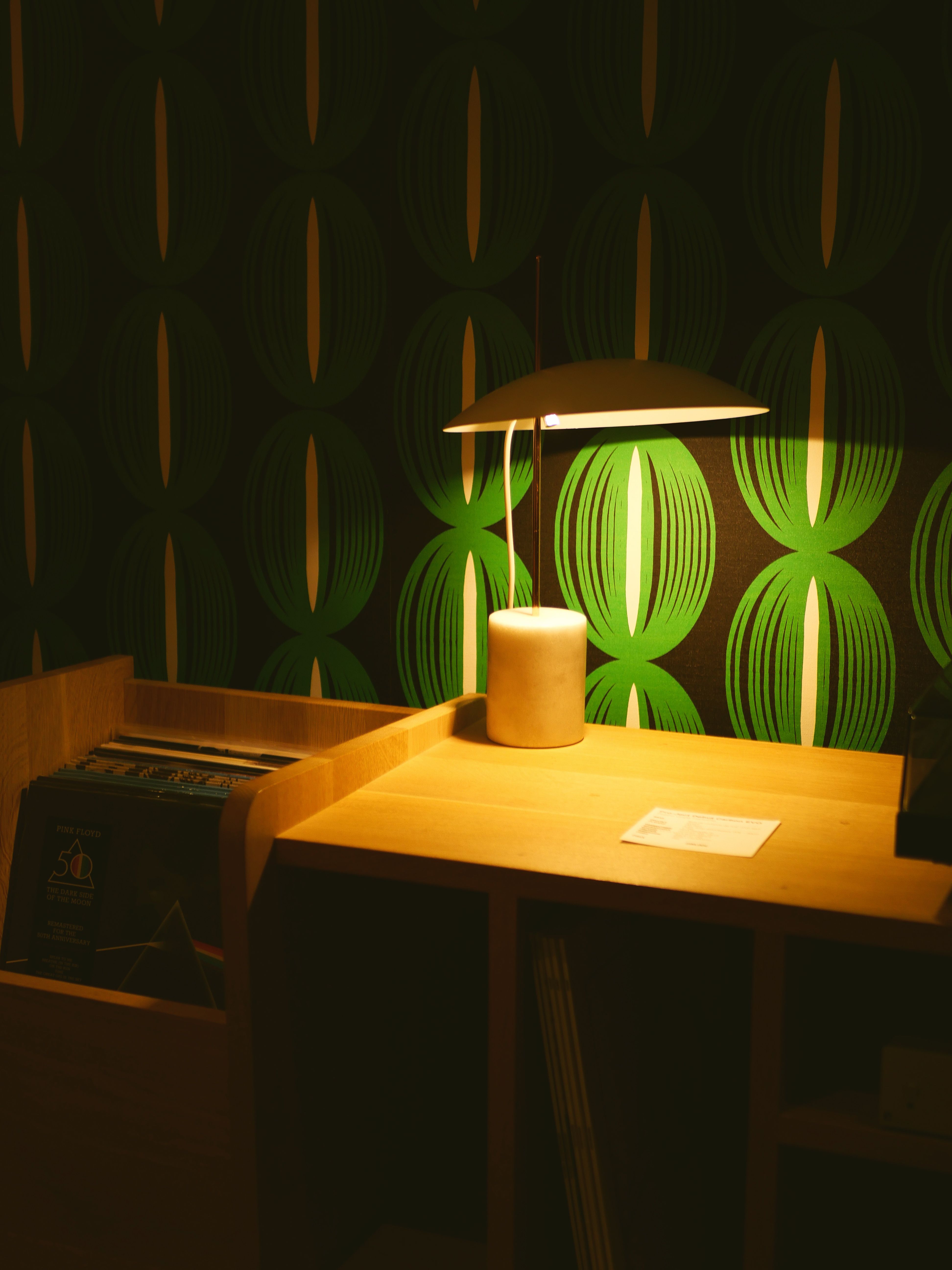 Warmly lit desk with lamp against a green vintage pattern wallpaper, perfect for cosy interior designs.