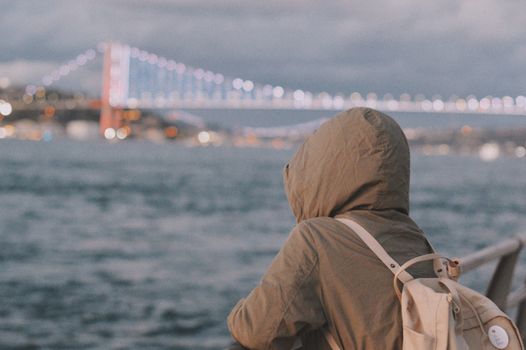 A person in a hooded jacket and backpack gazing at a lit bridge over a coastal view.