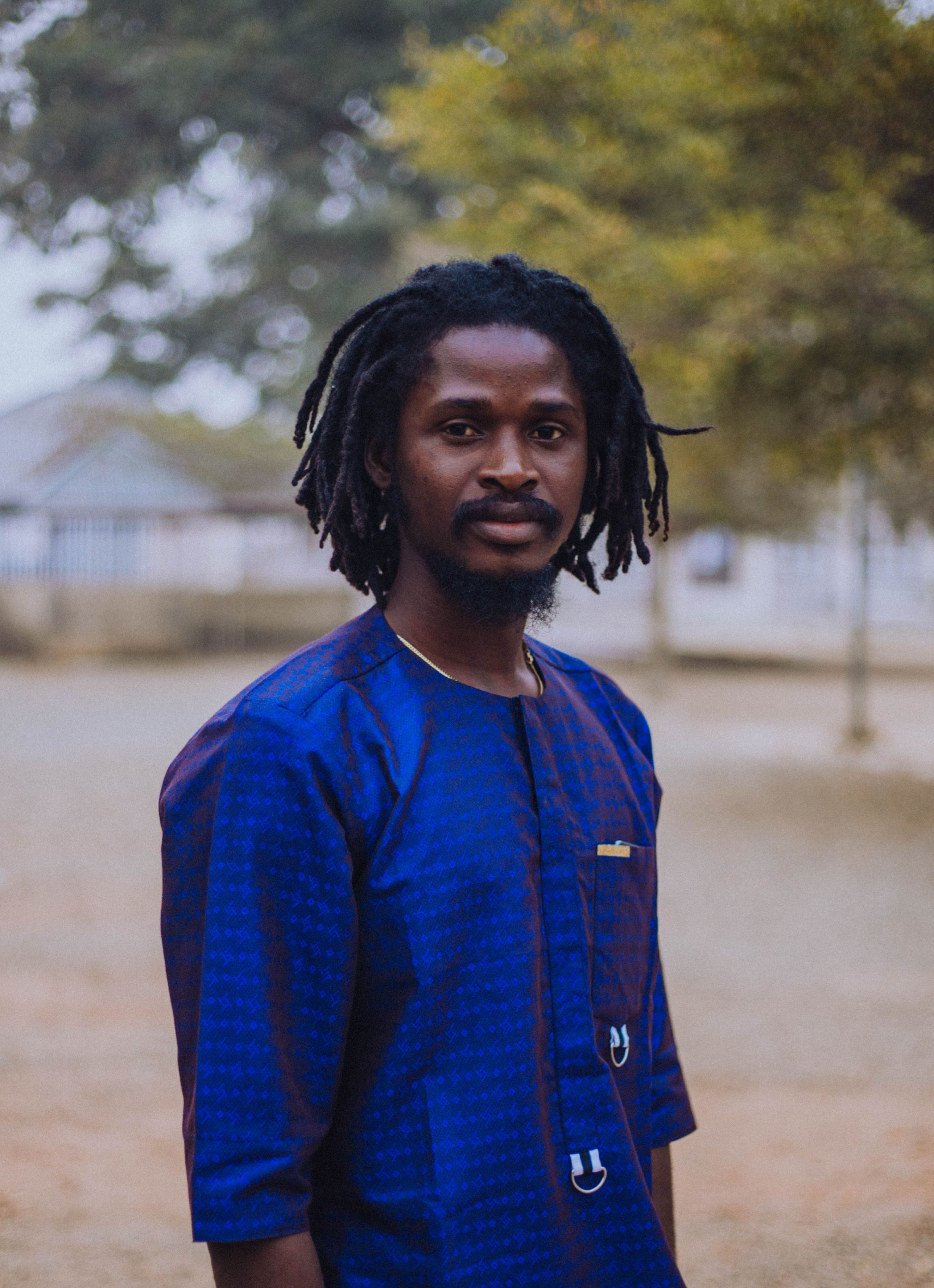 Portrait of a man in traditional Nigerian attire standing outdoors with trees in the background.