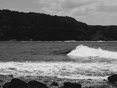 Moody black and white photo capturing ocean waves crashing near a cliff under a clouded sky.