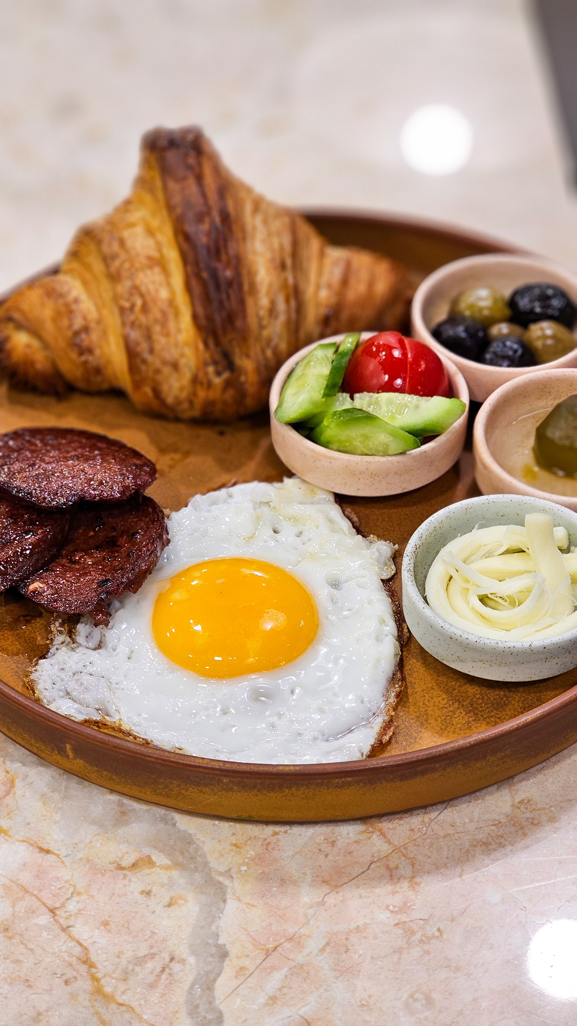 Appetizing breakfast platter featuring a fried egg, croissant, and fresh vegetables on a stylish wooden serving board.