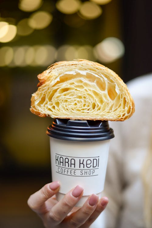 Free Close-up of a Person Holding a Paper Coffee Cup with a Croissant Sitting on Top of the Lid  Stock Photo