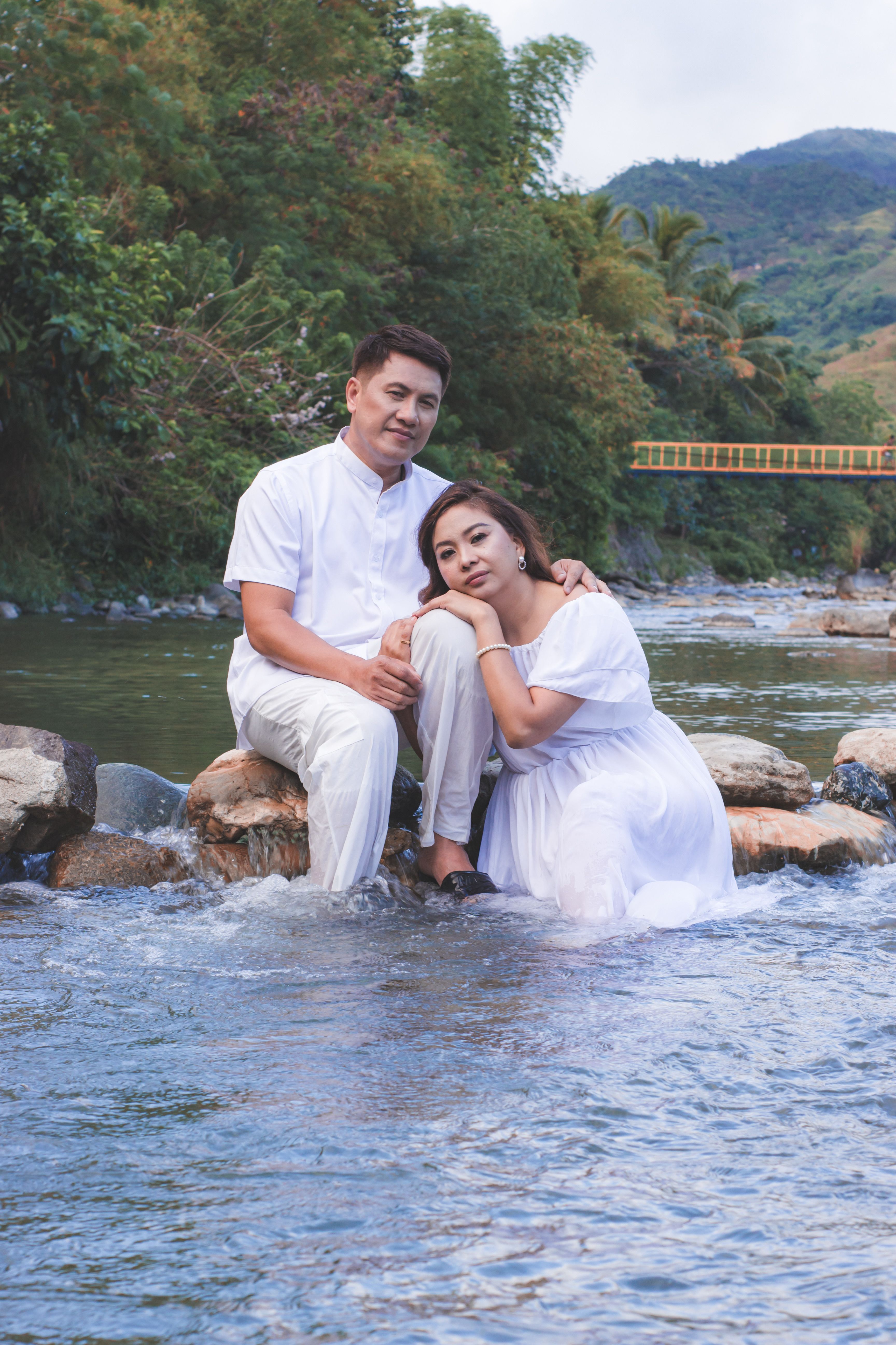 A couple in white attire sitting on rocks by a scenic river, surrounded by lush greenery.