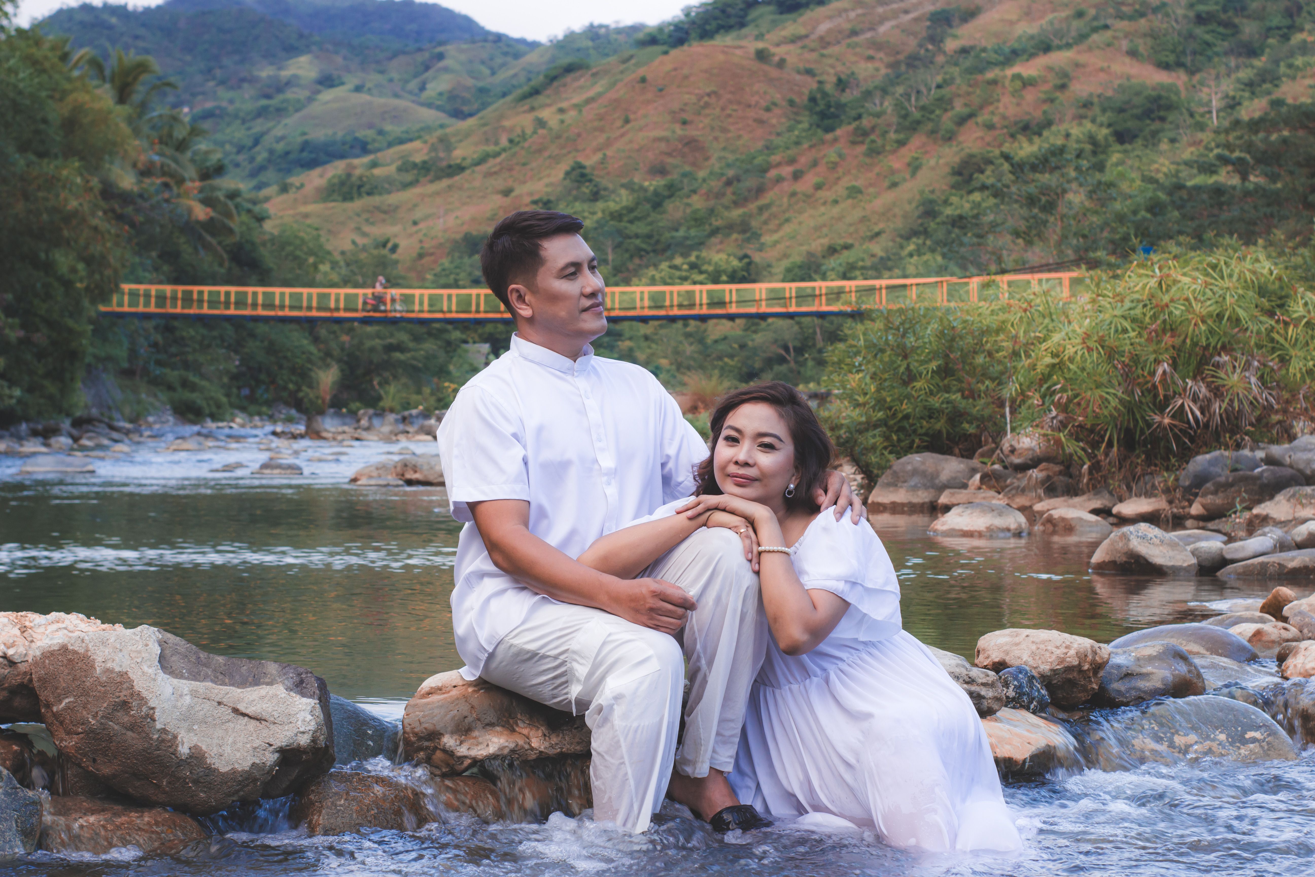 A loving couple embracing by a tranquil river, surrounded by lush hills and a scenic bridge.