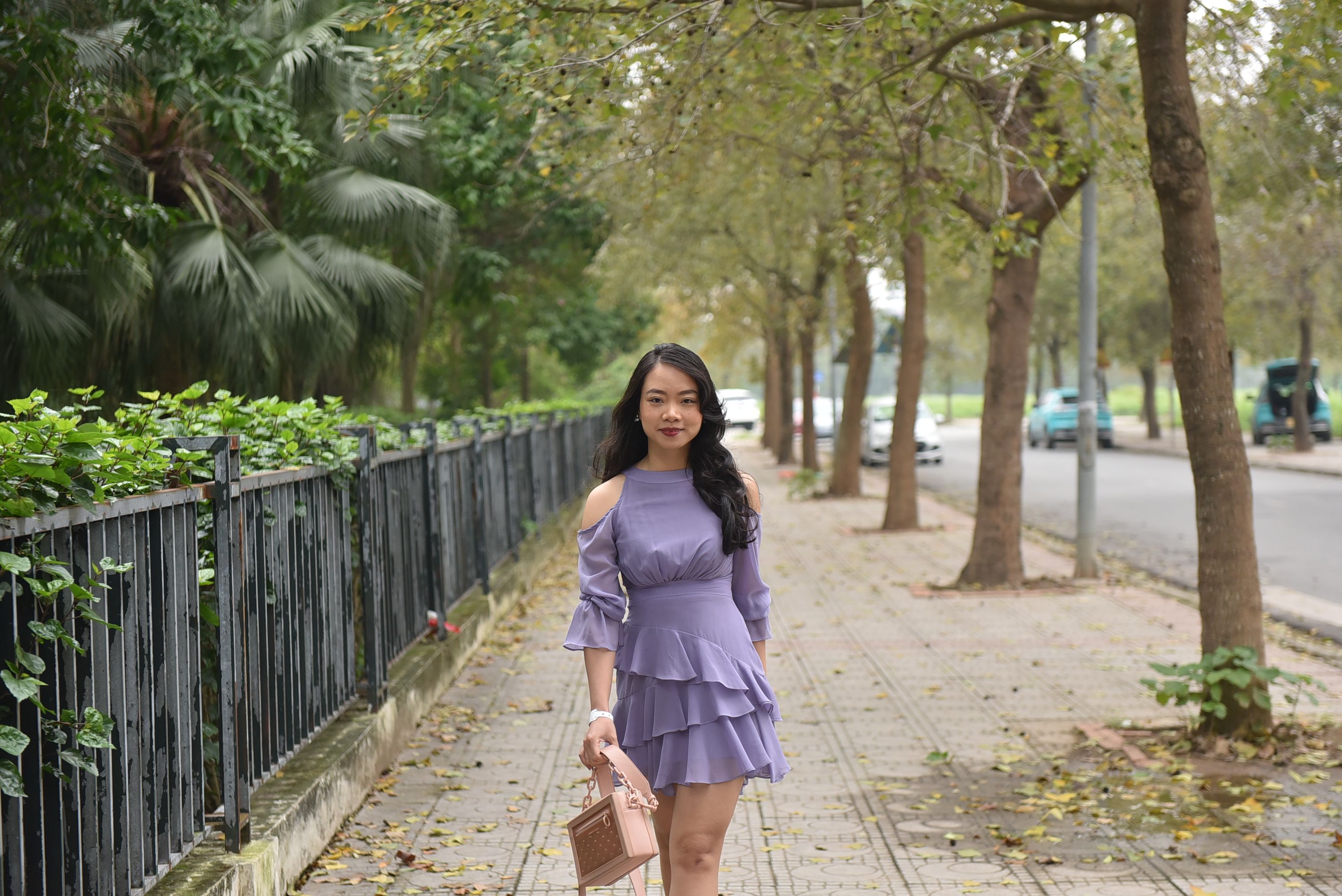 Stylish woman in purple dress walking along an urban sidewalk lined with trees.