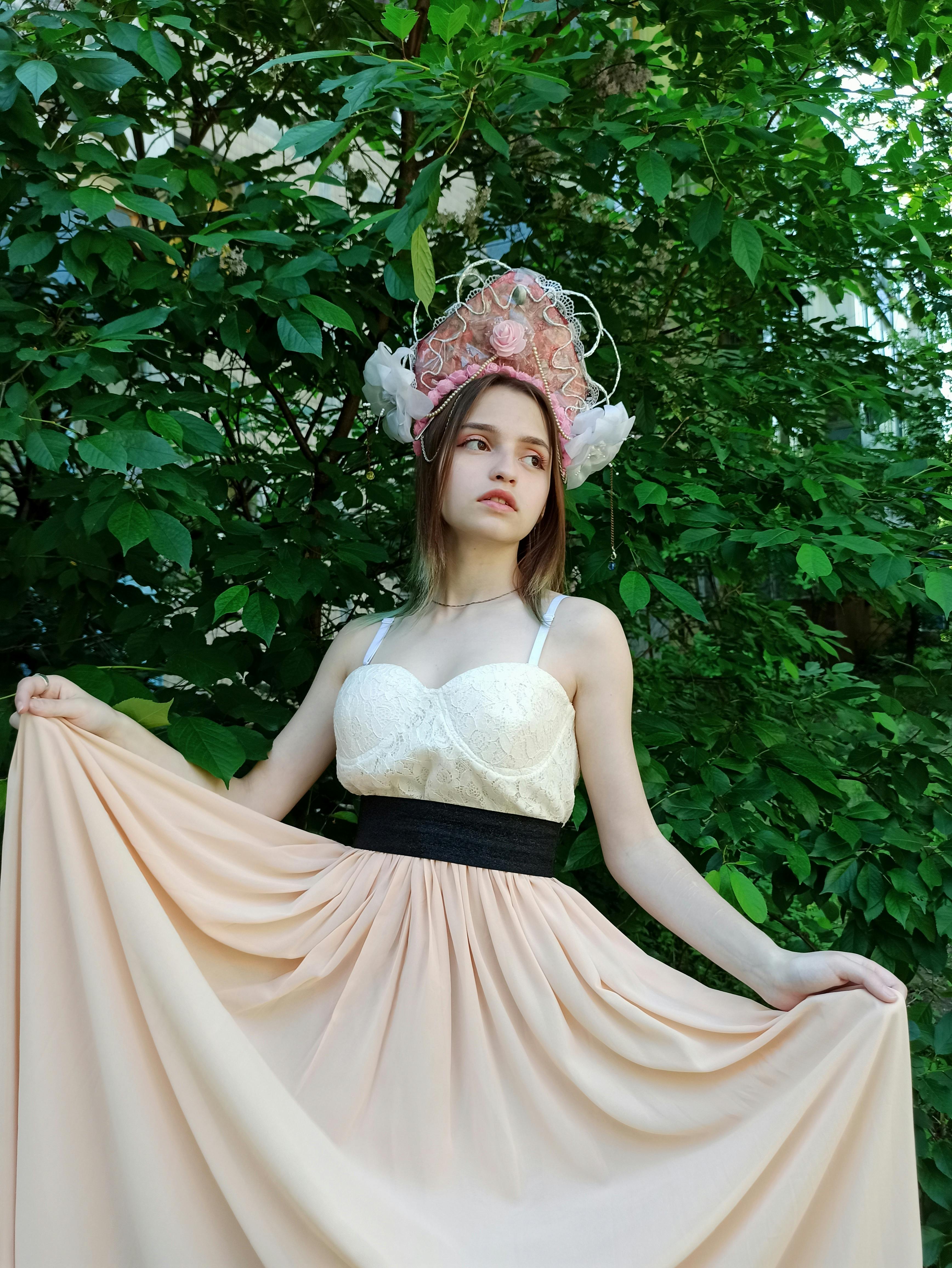 A beautiful woman in a flowing dress and kokoshnik stands gracefully among green foliage.