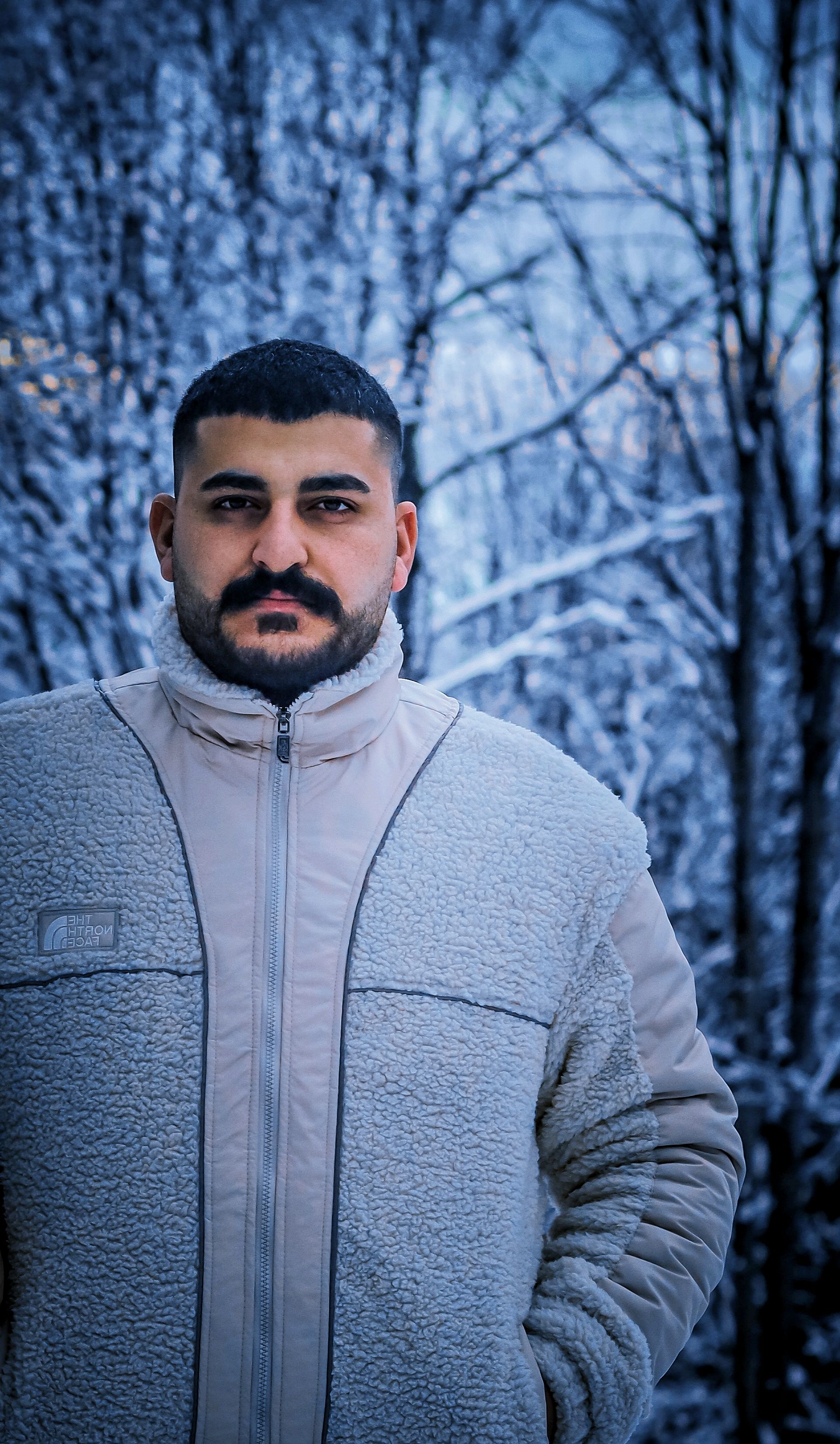 A man with a mustache in a warm jacket stands in a snowy winter forest.