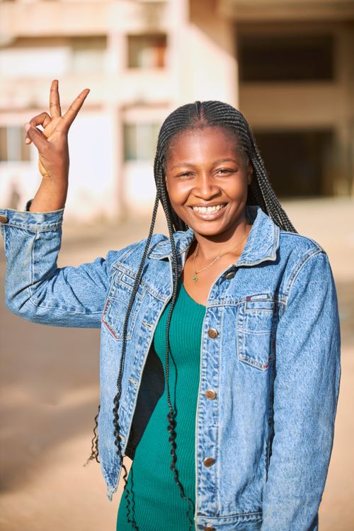 Free Portrait of a smiling woman in a denim jacket, posing outdoors with a peace sign. Stock Photo