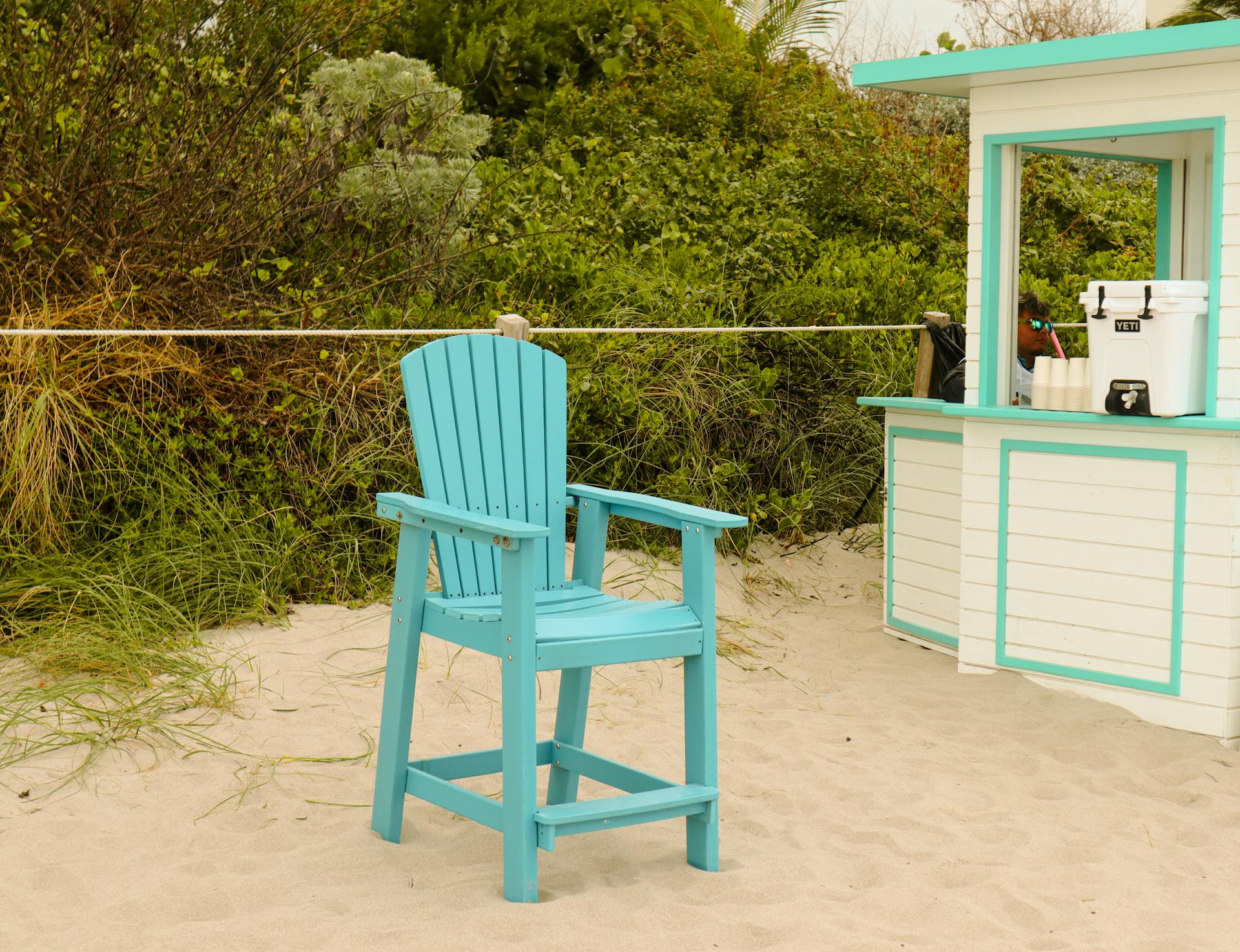 A bright turquoise beach chair on sandy shore by a seaside kiosk in summer.