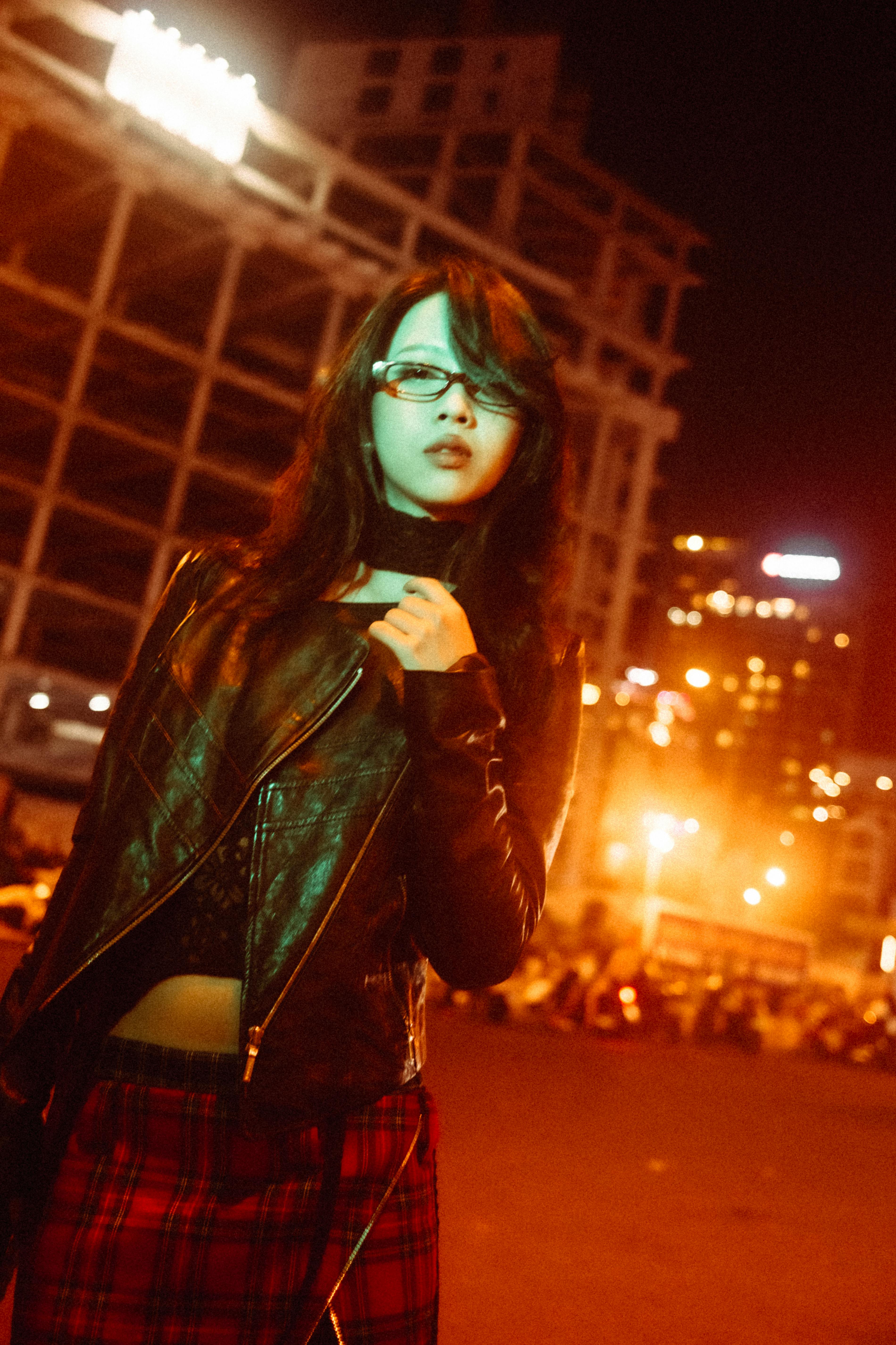 Stylish woman in leather jacket at night, city lights in the background.
