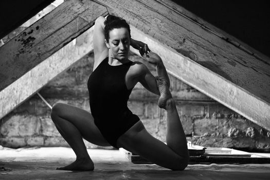 Black and white photo of a gymnast in a bodysuit stretching gracefully in an attic.