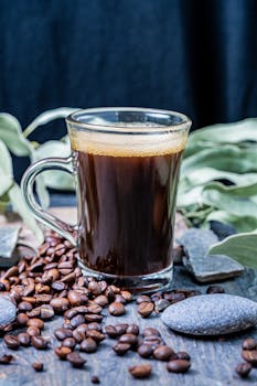 A clear glass cup filled with fresh steaming coffee surrounded by coffee beans and pebbles.