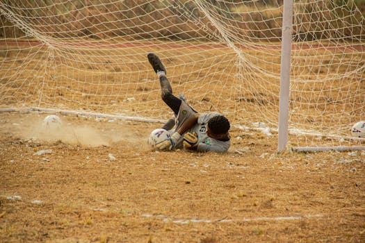 Energetic goalkeeper diving to save a soccer ball on a grassy pitch.