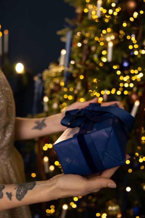 Free Close-up of a blue gift held by tattooed hands against a sparkling Christmas tree backdrop. Stock Photo