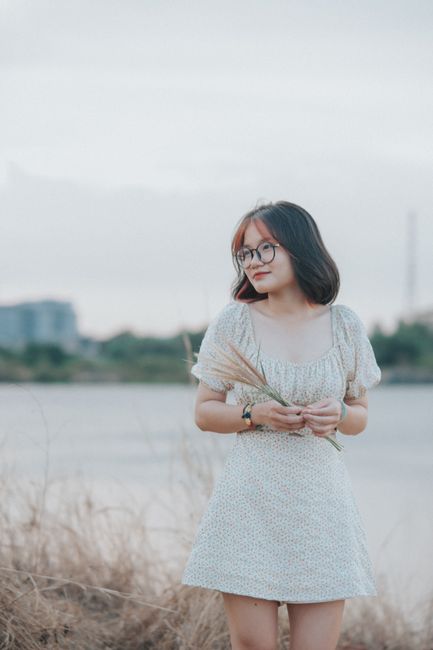 Asian woman in sundress holding plant by a river, showcasing serene rural fashion.