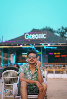 A fashionable man in sunglasses sits at a lively beach lounge, embodying summer vibes.