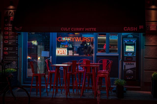 Charming night shot of a Berlin currywurst restaurant with vibrant neon sign and red chairs.