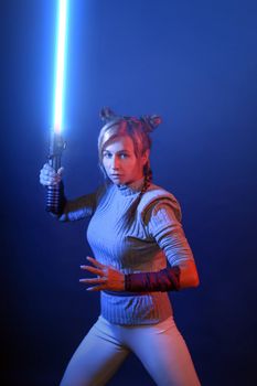 A woman in cosplay poses with a blue lightsaber against a dark background.