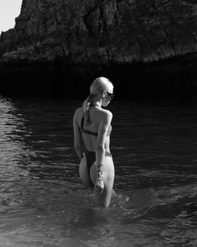 Stylish grayscale photo of a woman in a bikini posing at a rocky beach.