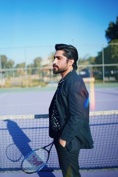 A fashionable man with a beard poses with a tennis racket on an outdoor court.