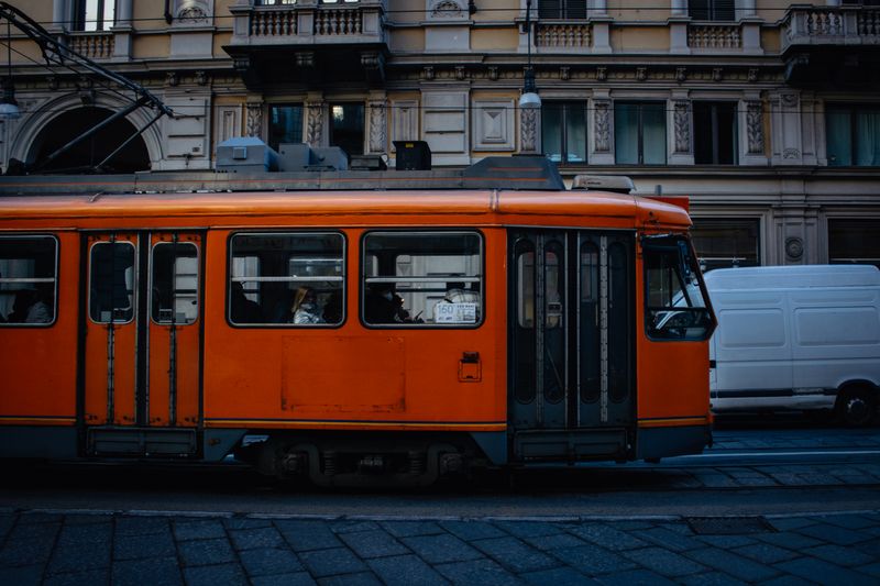 A striking orange tram travels through a bustling city street, showcasing urban public transportation.