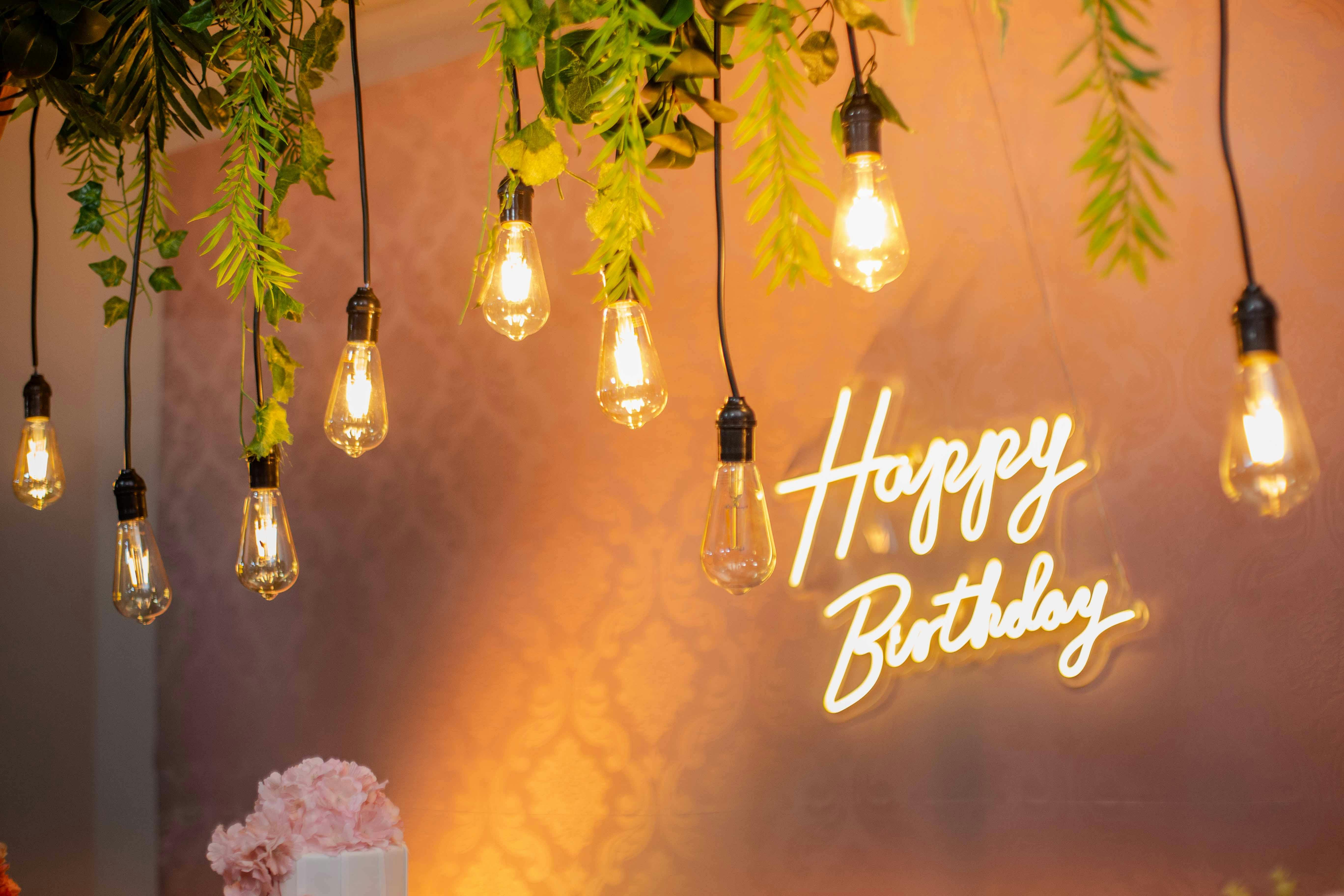 Stylish birthday setup featuring neon 'Happy Birthday' sign, hanging bulbs, and greenery for a festive ambiance.