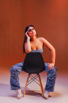 Fashionable young woman sitting on a chair, showcasing a trendy denim look in a studio setting.