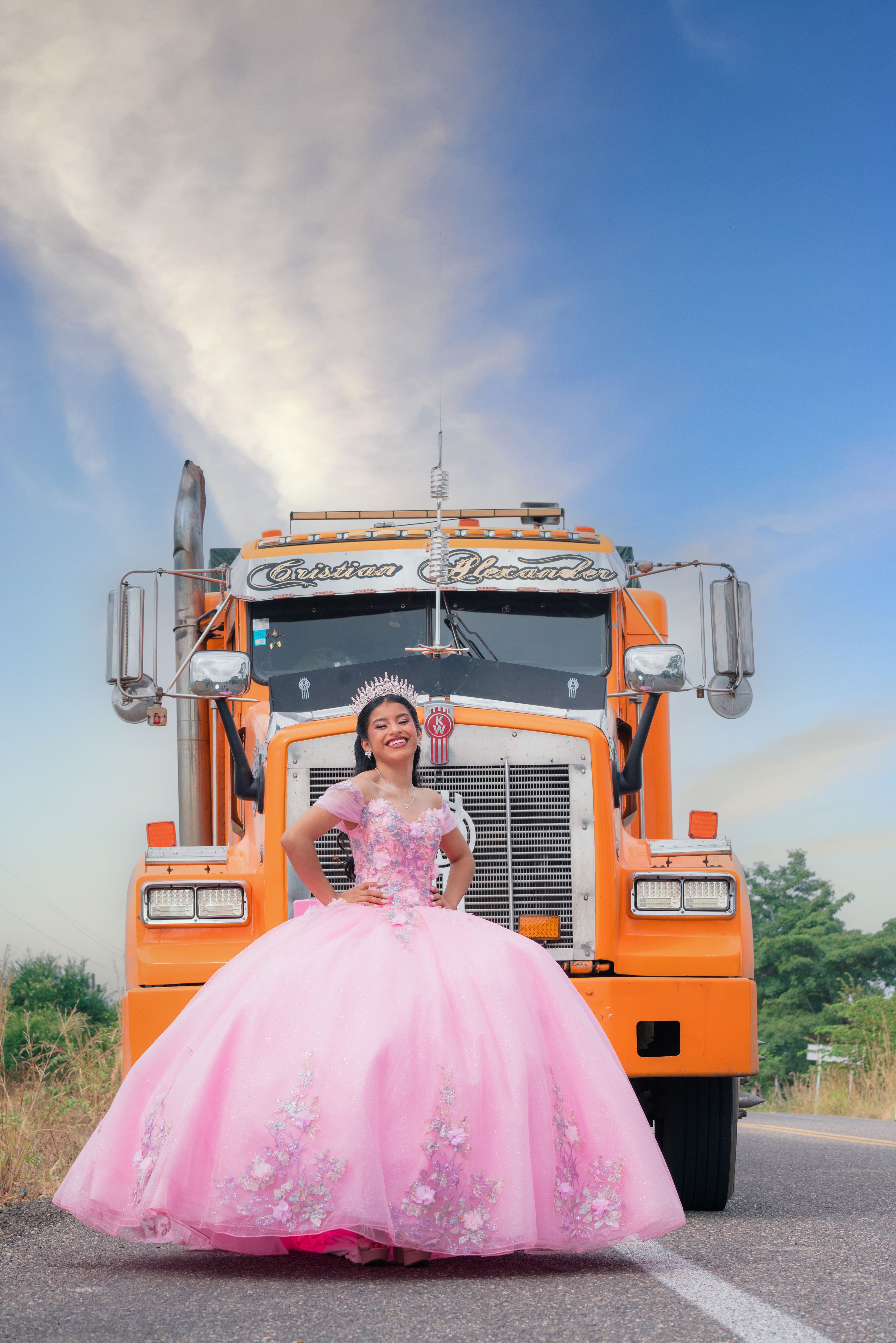 A young woman in a pink gown smiles in front of a vibrant orange truck under a clear blue sky.