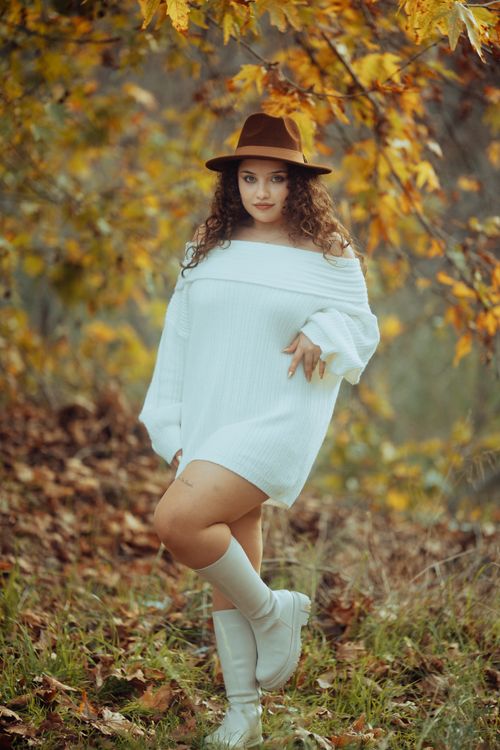 Free A fashionable woman in a white dress and hat poses outdoors amidst autumn leaves. Stock Photo