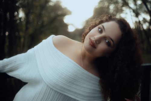 Free A woman in a white off-shoulder sweater leans, set against a natural forest background. Stock Photo