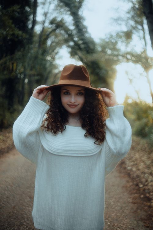 Free Young woman with curly hair wearing a brown fedora and white sweater, posing outdoors on a forest path. Stock Photo
