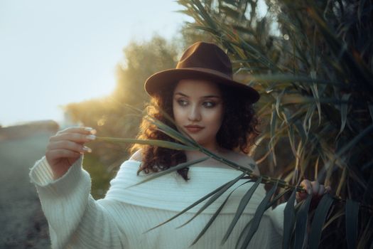 A young woman in a white sweater and brown fedora stands amidst sunlit greenery, exuding fashion and elegance.