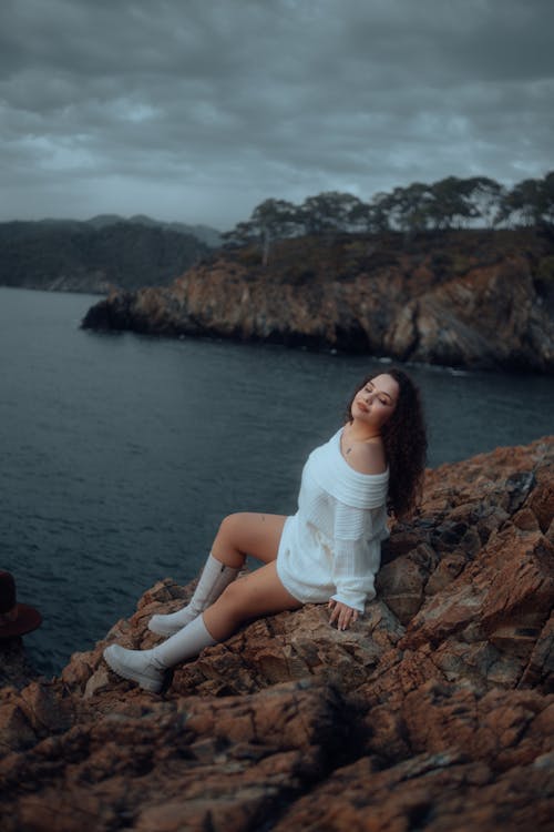 Portrait of a Pretty Brunette Wearing a White Sweater Sitting on Coastal Rocks