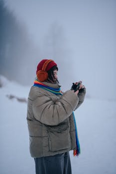 A woman in winter clothes captures a snowy scene with her smartphone in a foggy, serene landscape.