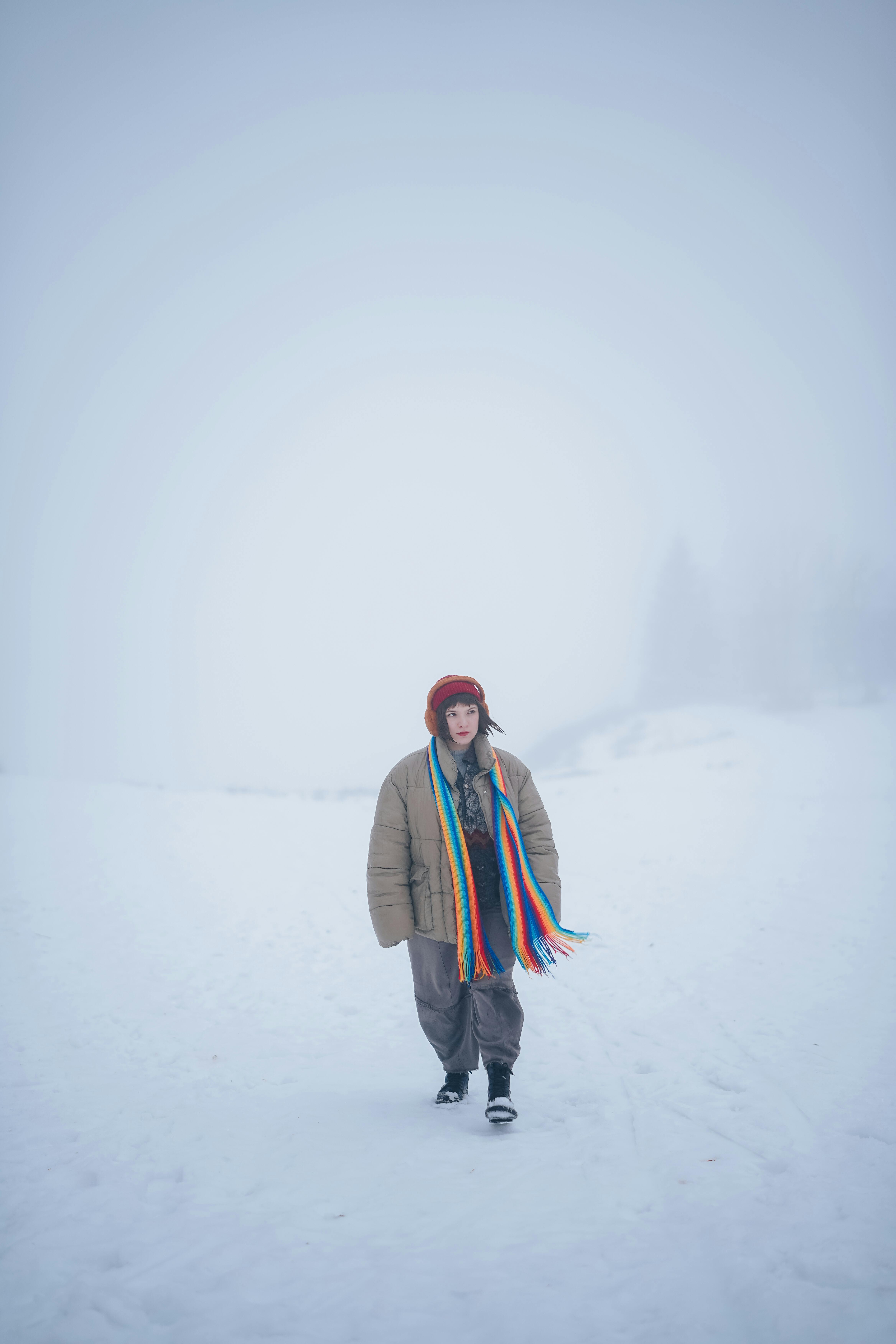 A woman walks through a snowy landscape, wearing a colorful scarf and winter clothing, creating a vivid contrast against the muted background.