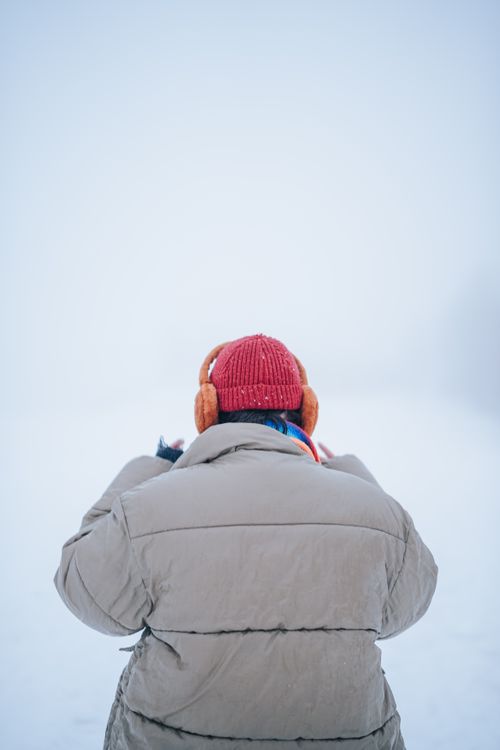 Free Back view of a person in winter attire, earmuffs, and red hat standing in a snowy scenery. Stock Photo
