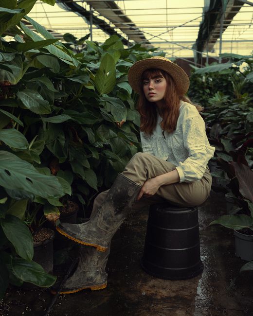 Young woman in straw hat sitting among lush greenery in a greenhouse.