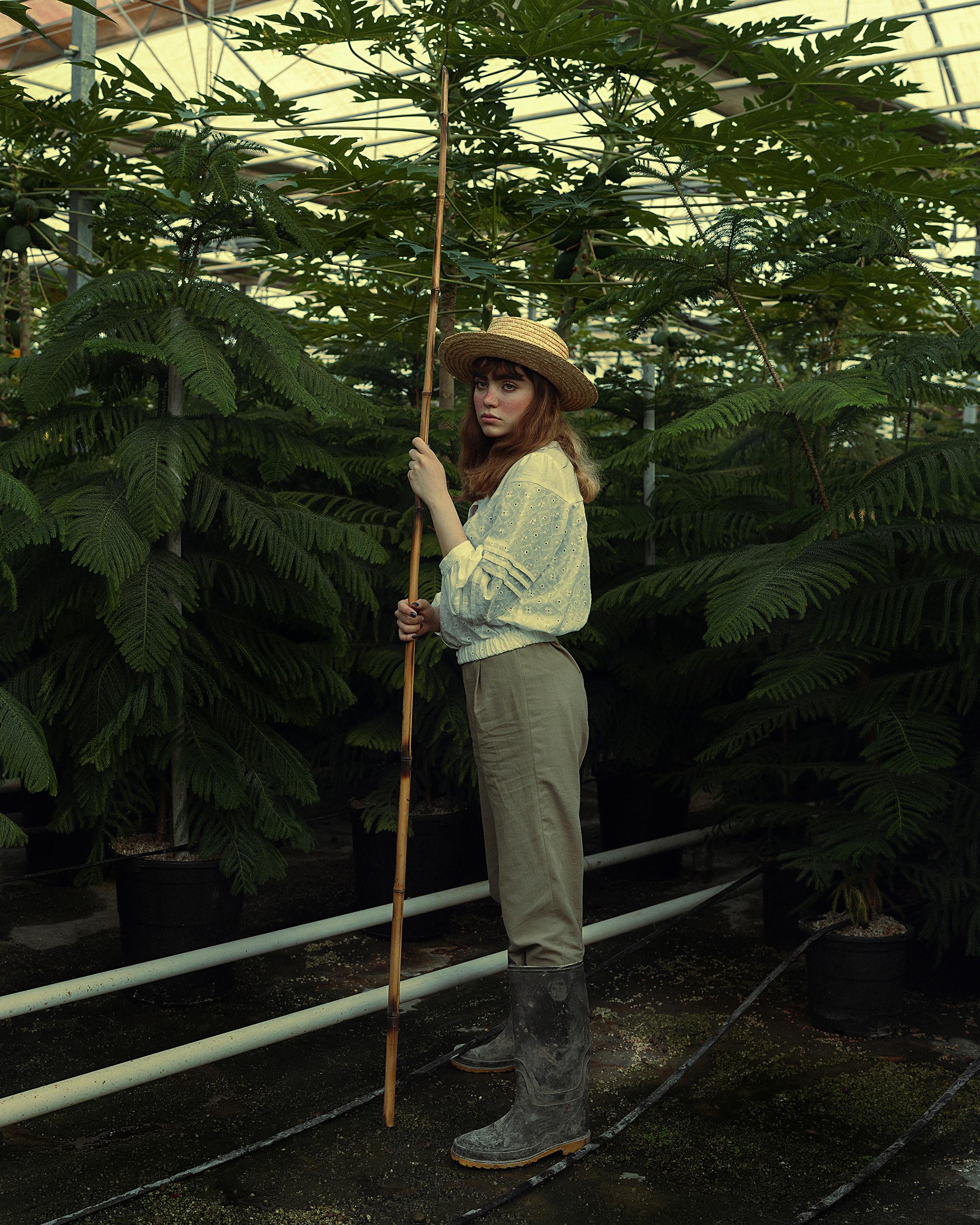 A young woman in a hat tends plants with a bamboo stick in a greenhouse, wearing boots and a white shirt.