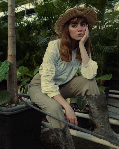 Free Fashionably dressed woman in a greenhouse, deep in thought, wearing a straw hat and white shirt. Stock Photo