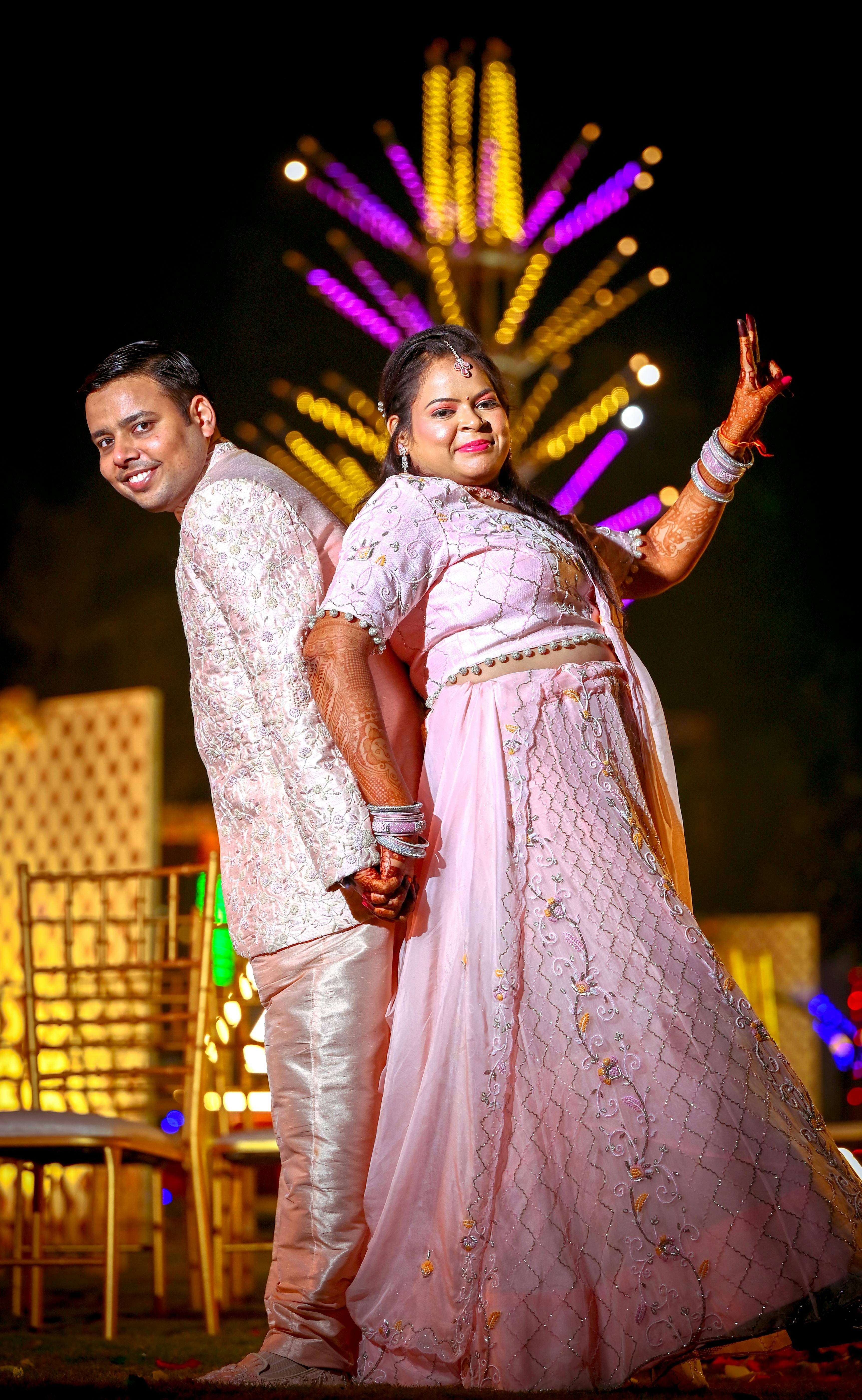 A joyful Indian couple dressed in traditional attire at a vibrant night event with colorful lights.