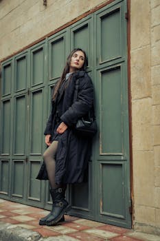 Fashionable woman in a black coat leans against a rustic green door, showcasing urban style.