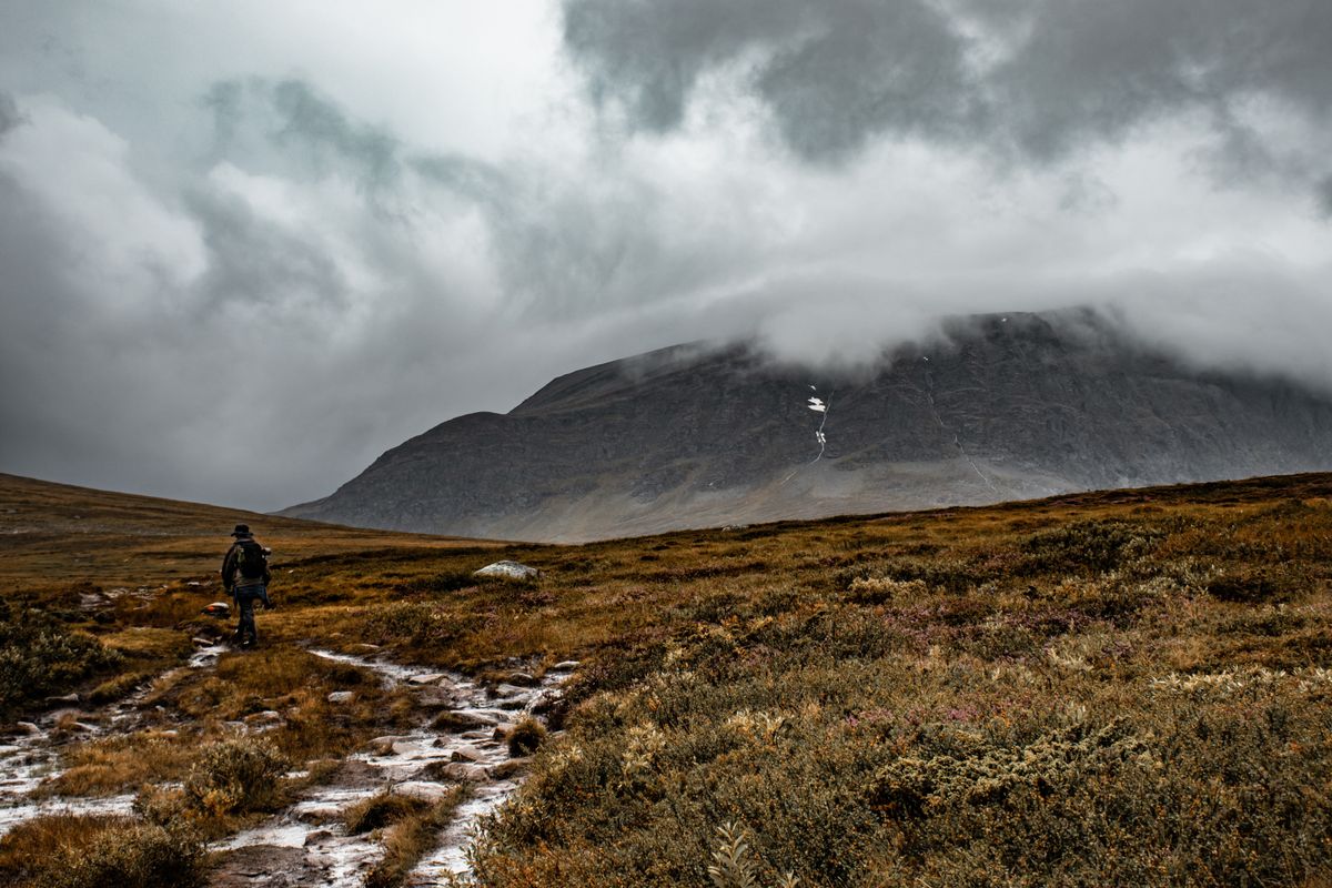 A single hiker on a wet, part-snowed trail across open highland, low storm cloud swallowing the peak ahead, visibility dropping by the step.