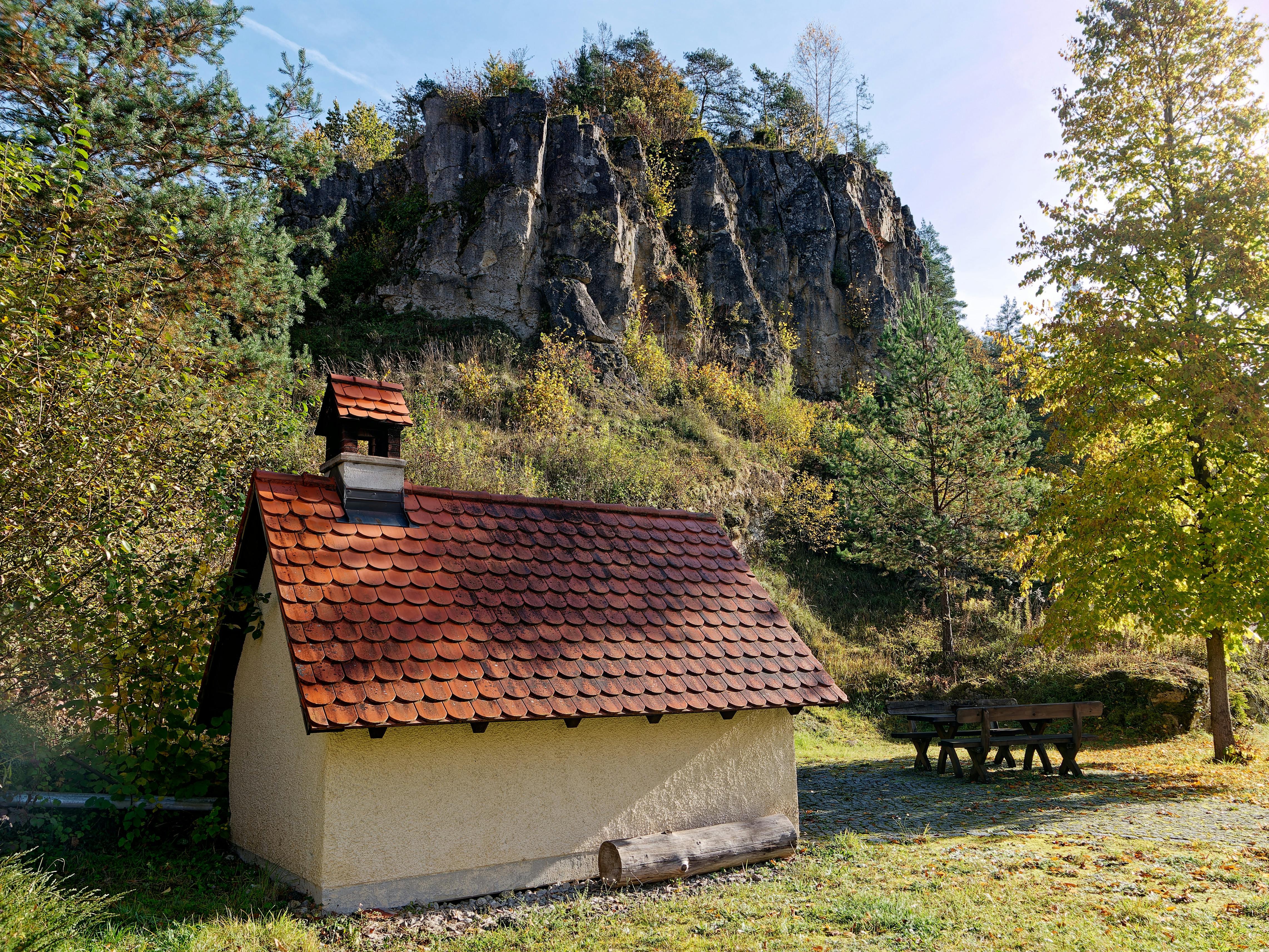 Rustic shed with a red roof nestled beside a rocky hill on a sunny day, surrounded by trees.