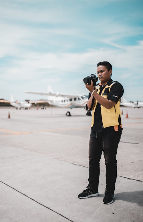 Pria Dengan Rompi Kuning Dan Kemeja Hitam Berdiri Di Landasan Pacu Memegang Kamera