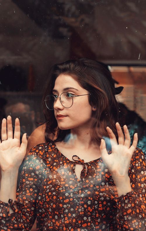 Free Portrait of a stylish young woman with eyeglasses behind a windowpane, captivating expression. Stock Photo