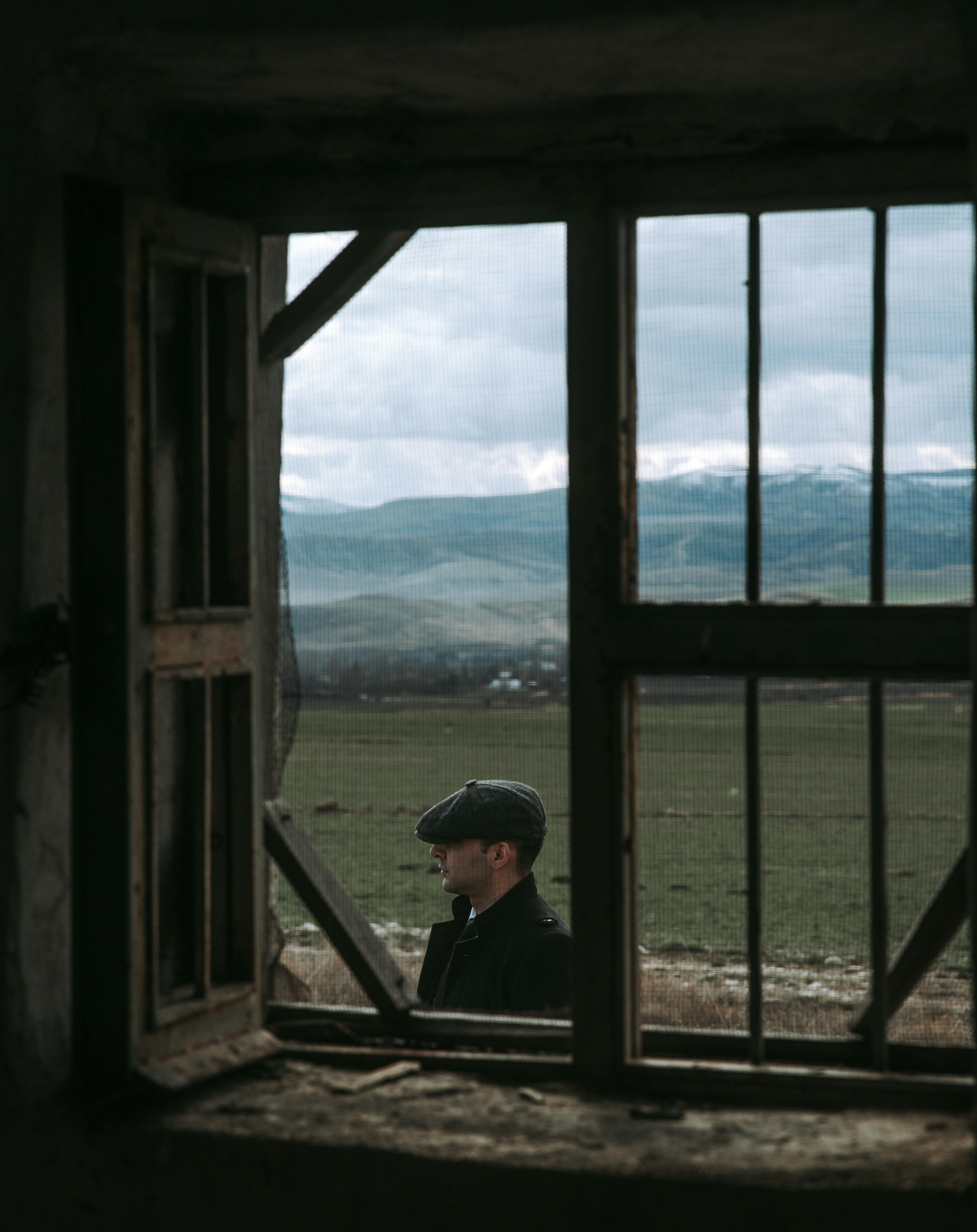 A man in a flat cap stands on a rural landscape viewed through a rustic window frame.