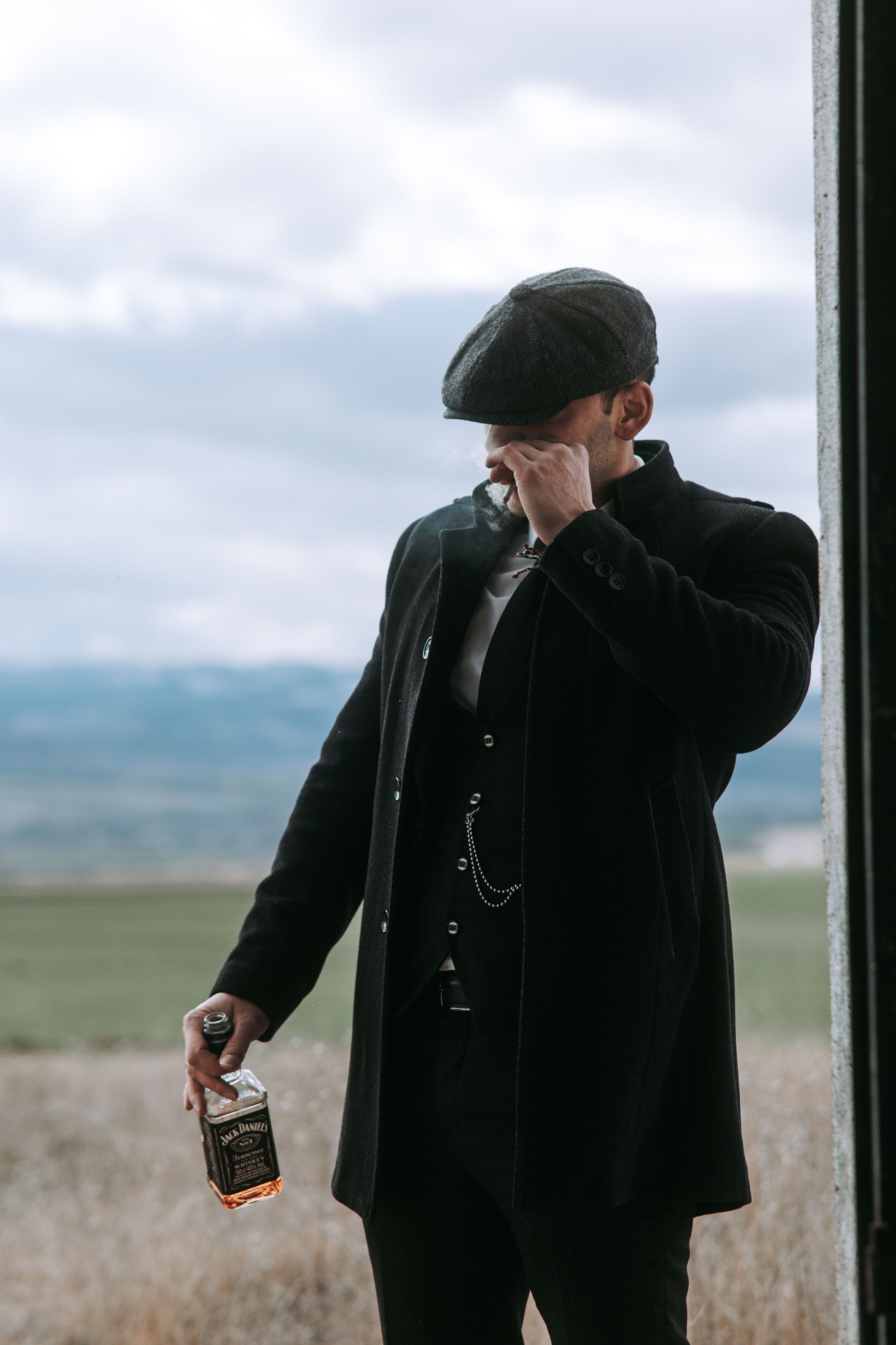 Man in elegant attire with whisky and cap in rural setting, blending fashion with leisure.