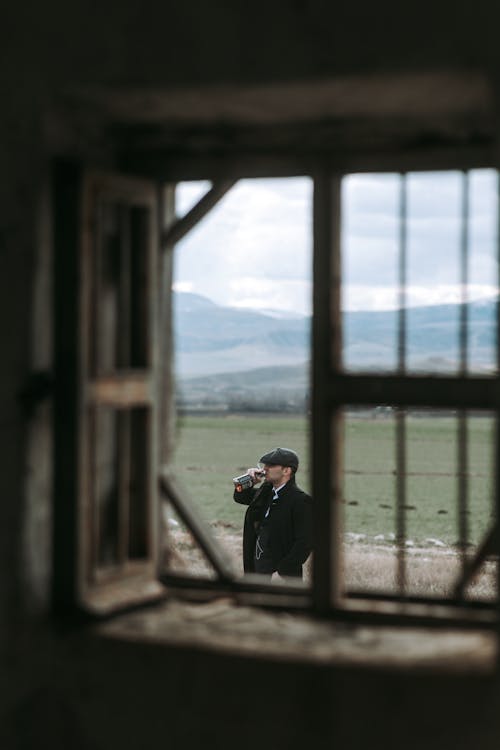 Man in a Flat Cap Drinking Whiskey Outside the Window of a Cottage