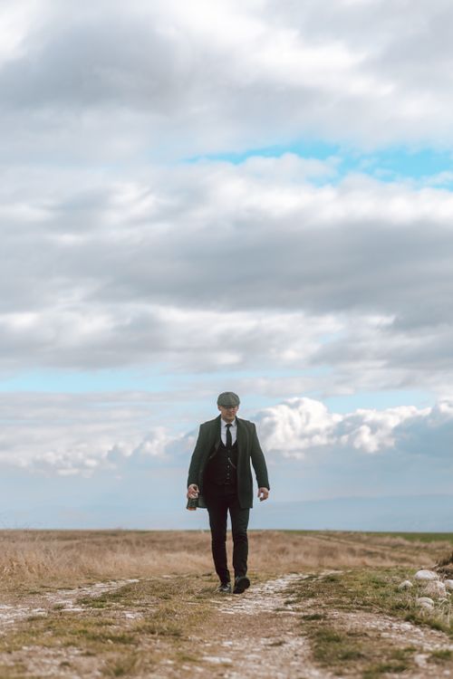 Free A well-dressed man in a flat cap walks along a remote path beneath a cloudy sky. Stock Photo