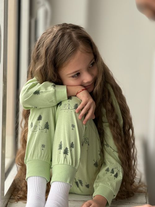 Free A young girl with long curly hair sits by a window, wearing green pajamas and daydreaming. Stock Photo